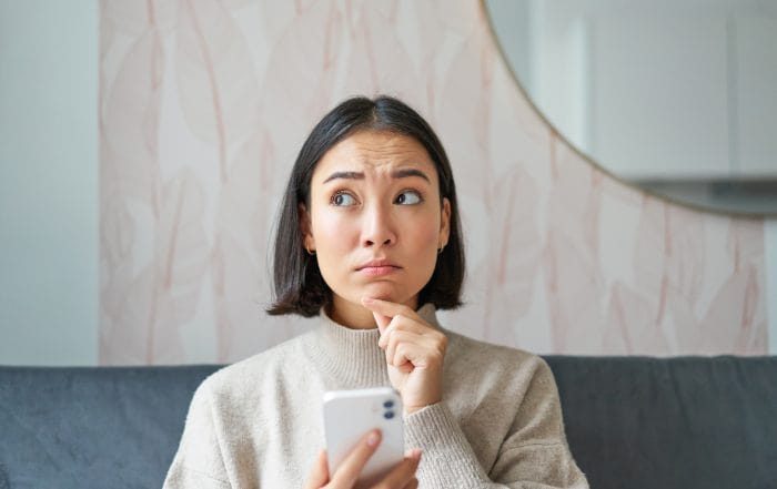Portrait of girl sitting on sofa with smartphone looking thoughtful and hesitant at mobile phone screen - Oasis Credit