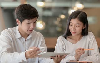 Two colleagues review a tablet together as the man uses a stylus and the woman holds an orange pencil.