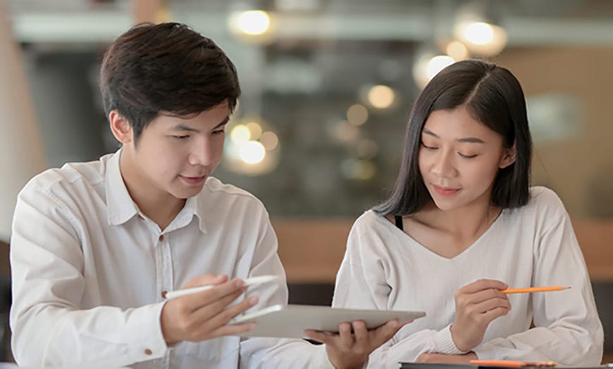 Two colleagues review a tablet together as the man uses a stylus and the woman holds an orange pencil.